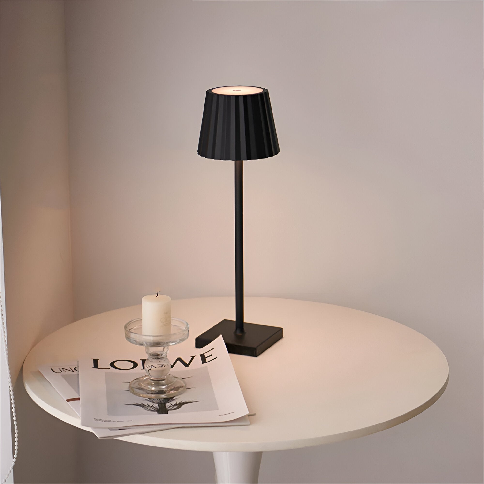 Black table lamp on a round white table with a candle and magazine against a gray wall.