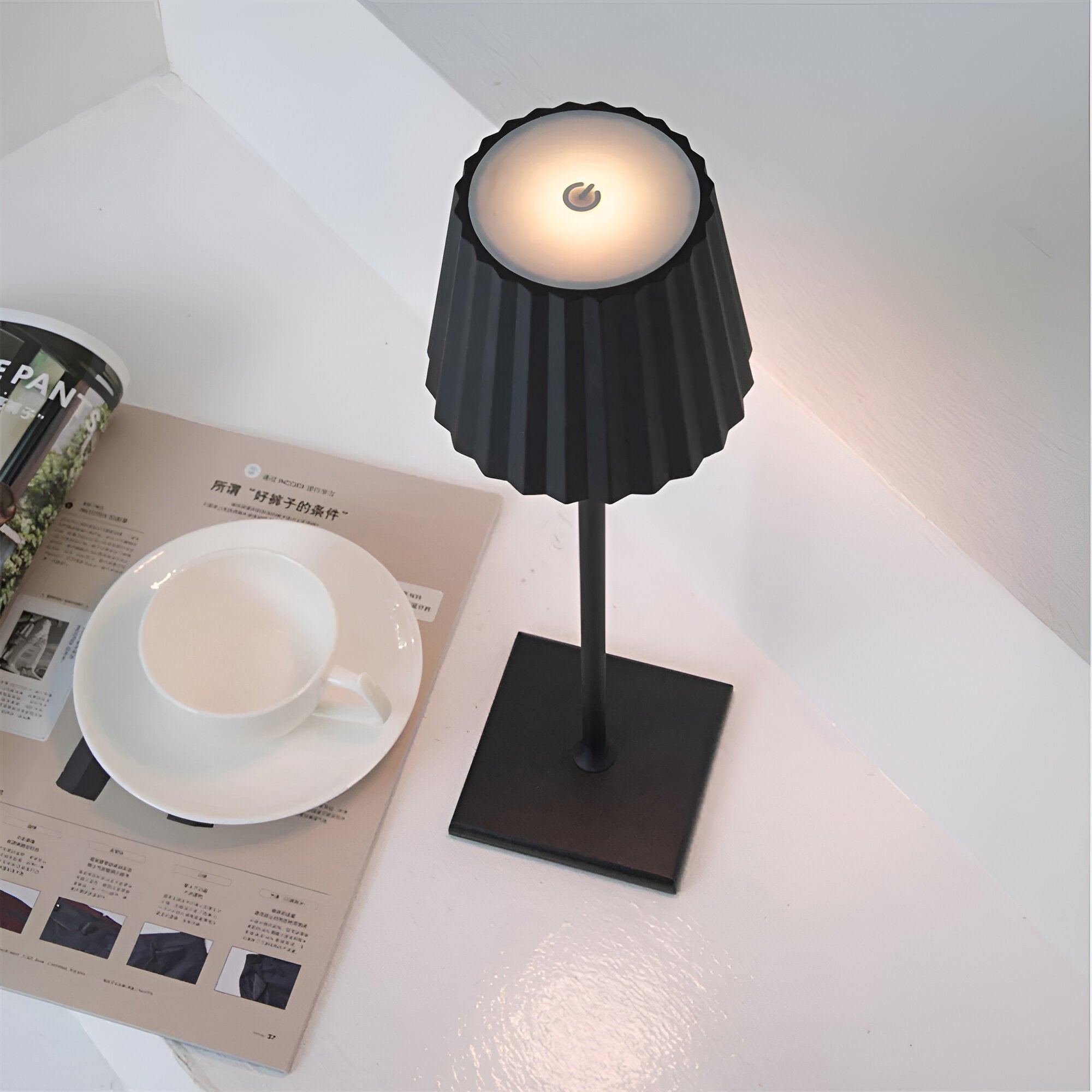 Black table lamp on a white surface with a magazine and cup in the background